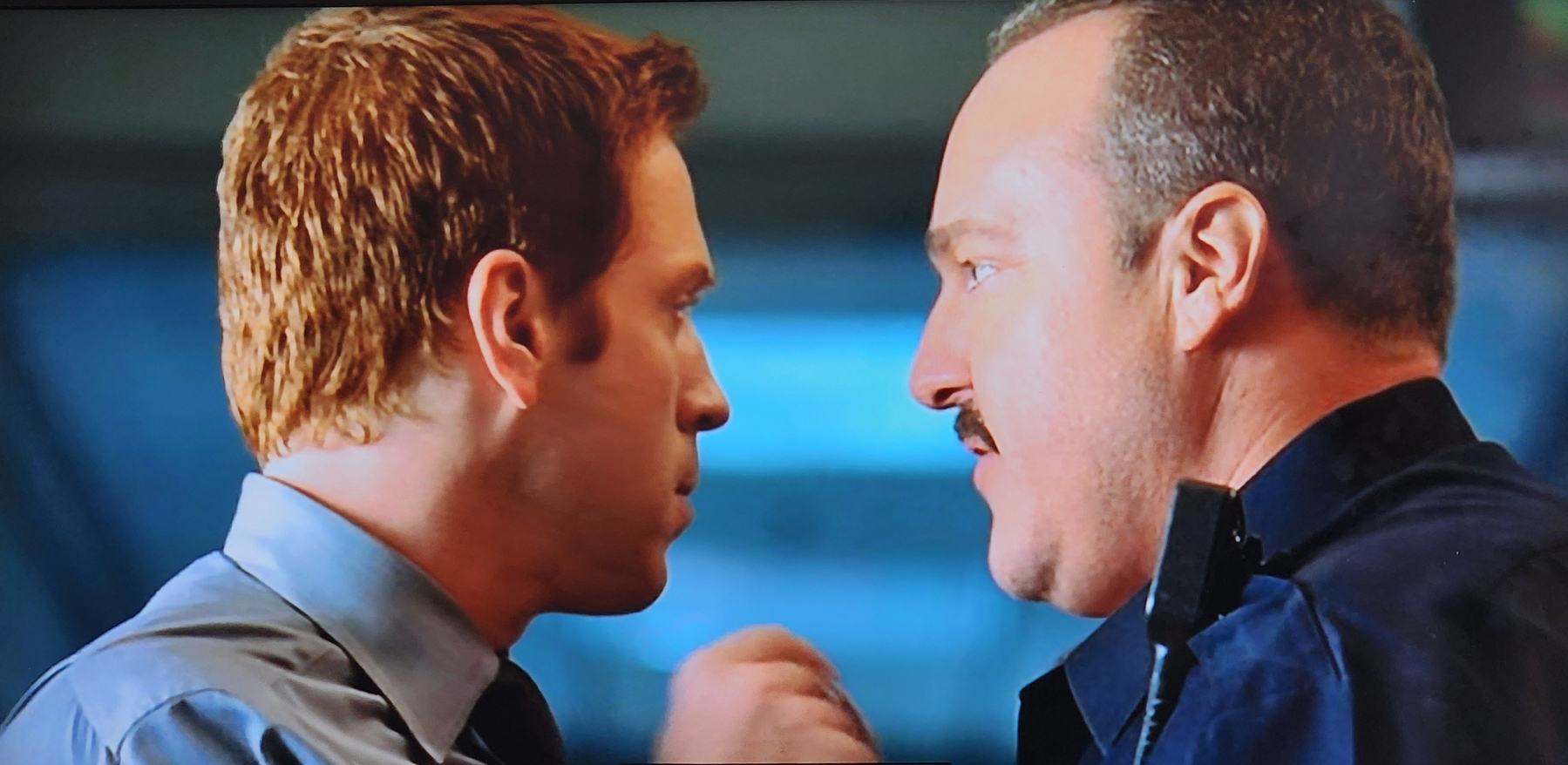 Two men, one with reddish hair and wearing a tie, and the other with short hair and a mustache, are facing each other closely, appearing to be in a tense conversation.